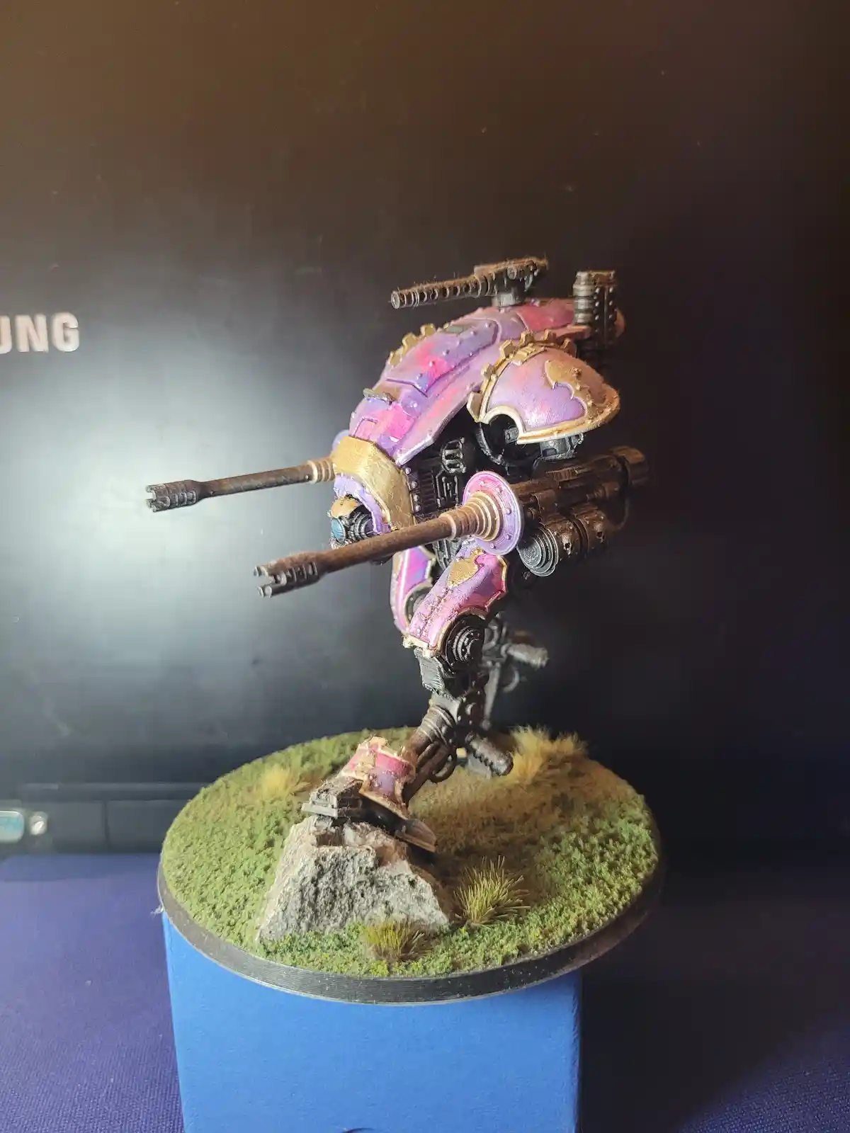 A heavily armored, purple and gold painted sci-fi war machine model stands on a circular base with green moss and a small…