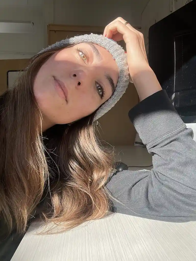 A young woman with long brown hair and a grey beanie looks directly at the camera with a relaxed expression while resting…