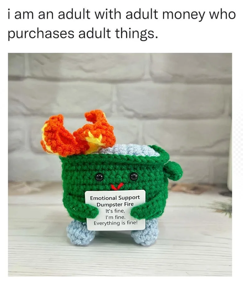 A green crocheted plush toy resembling a dumpster with orange flames on top, holding a sign that reads 'Emotional Support…