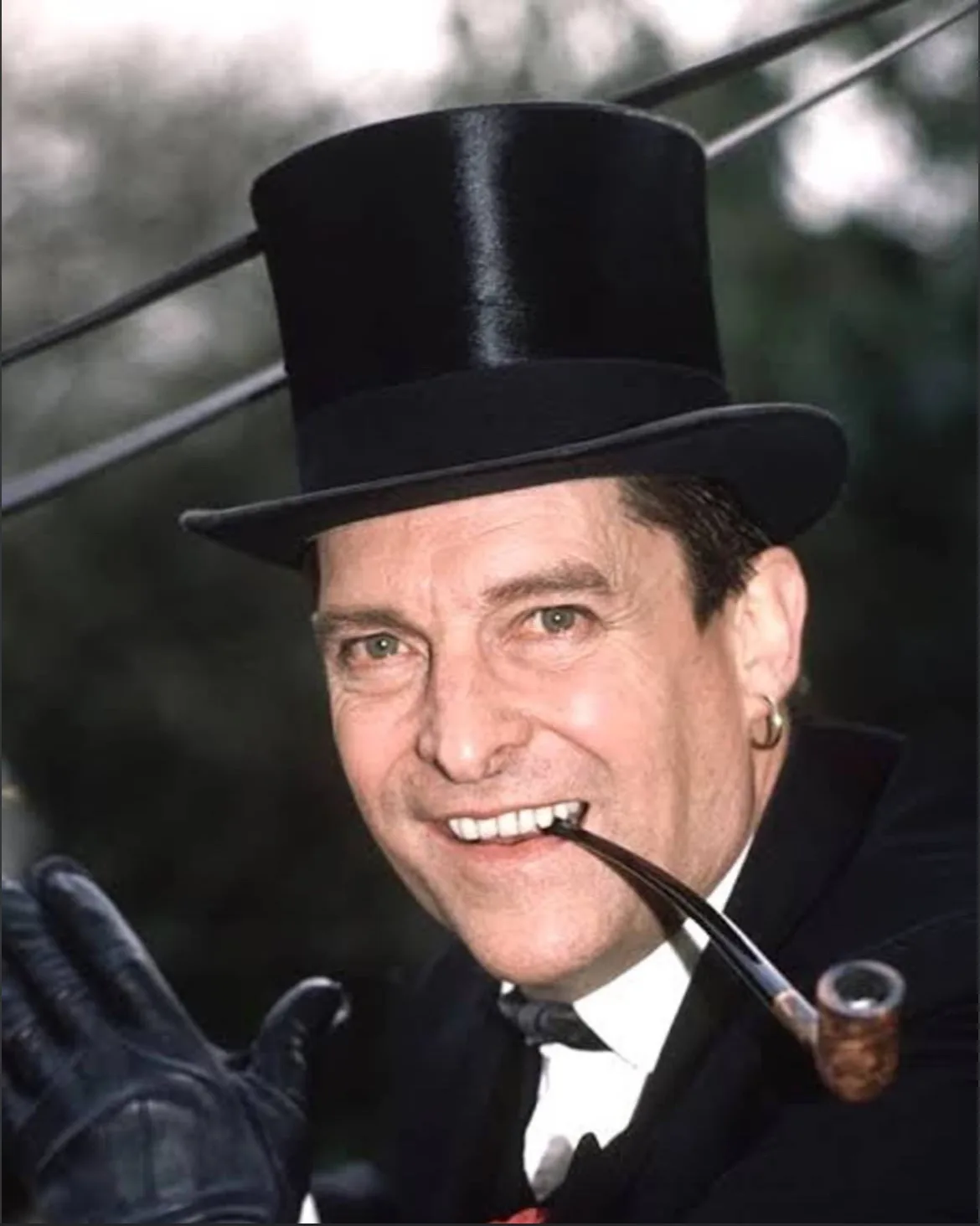 a man in a black top hat, black suit, white shirt, and black bow tie smiles while holding a pipe in his mouth and a gloved…