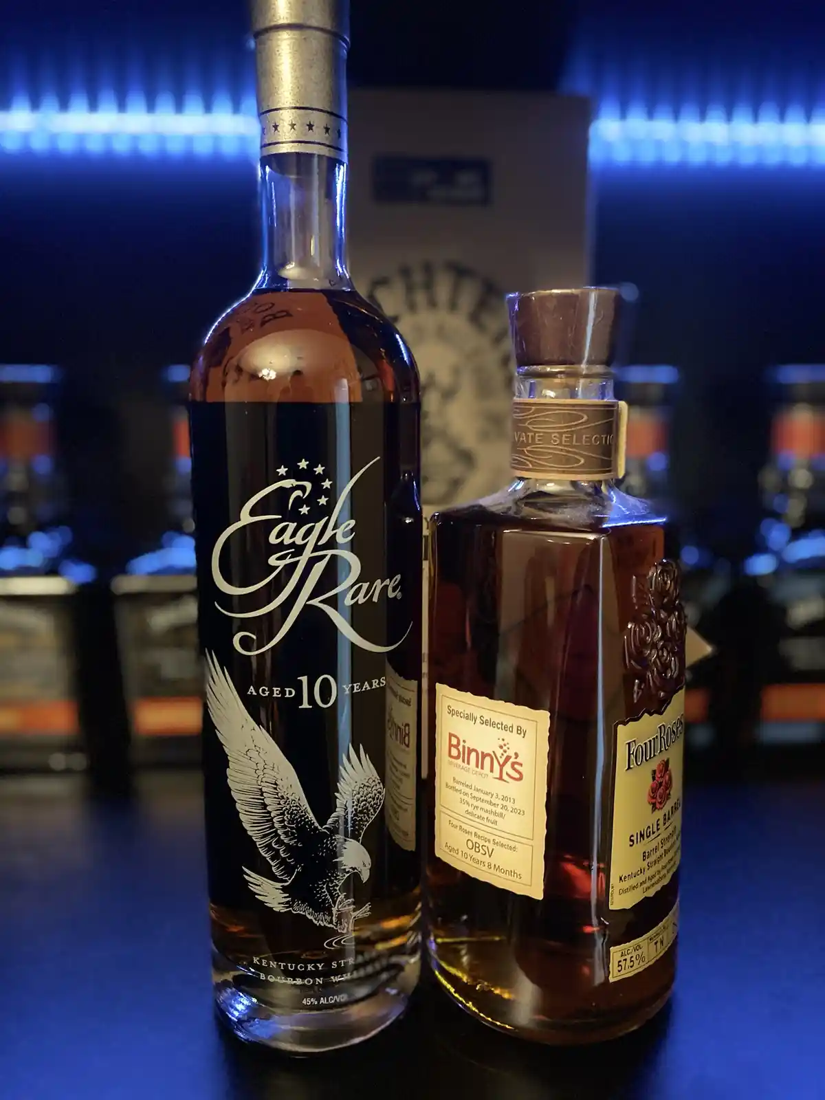 Two bourbon bottles are displayed side-by-side on a dark surface under blue ambient lighting. The taller bottle on the left…