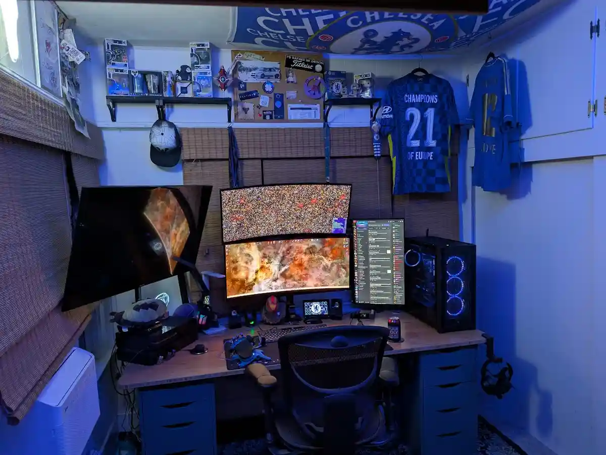 A dimly lit, blue-hued gaming room features a desk with three monitors displaying cosmic imagery, a gaming chair, and a PC…