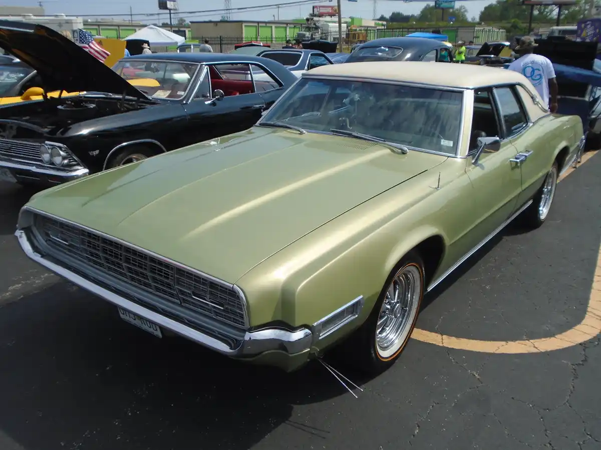 A light green classic American car with chrome trim and whitewall tires is parked at a car show, with other vintage cars and…
