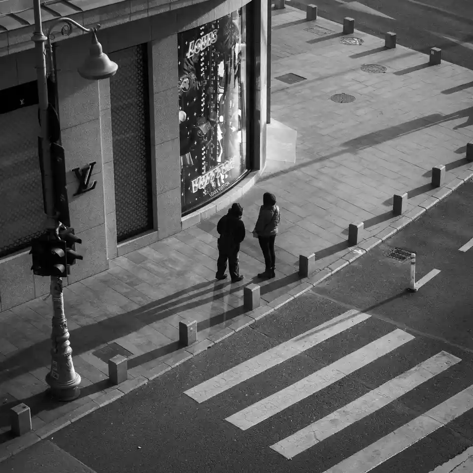 A high-angle black-and-white photo shows two people standing on a sidewalk in front of a Louis Vuitton store, looking at the…