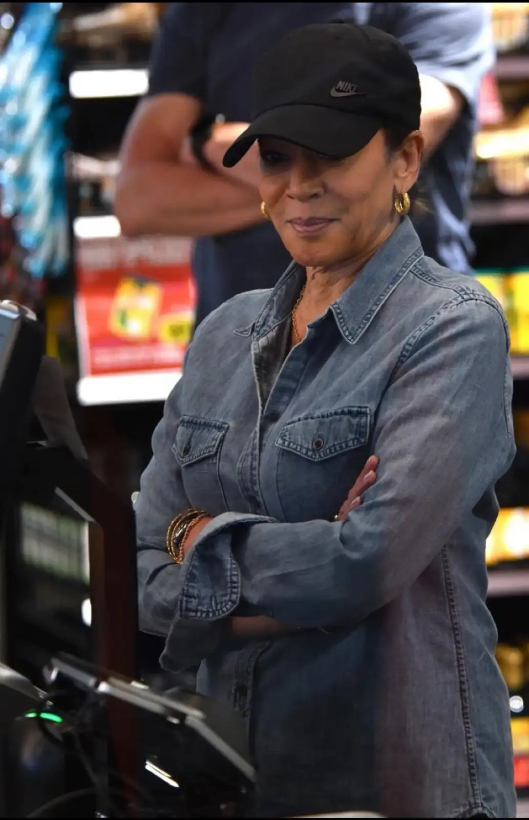 A woman wearing a black Nike cap and denim shirt stands with her arms crossed at a checkout counter, looking sternly at the…