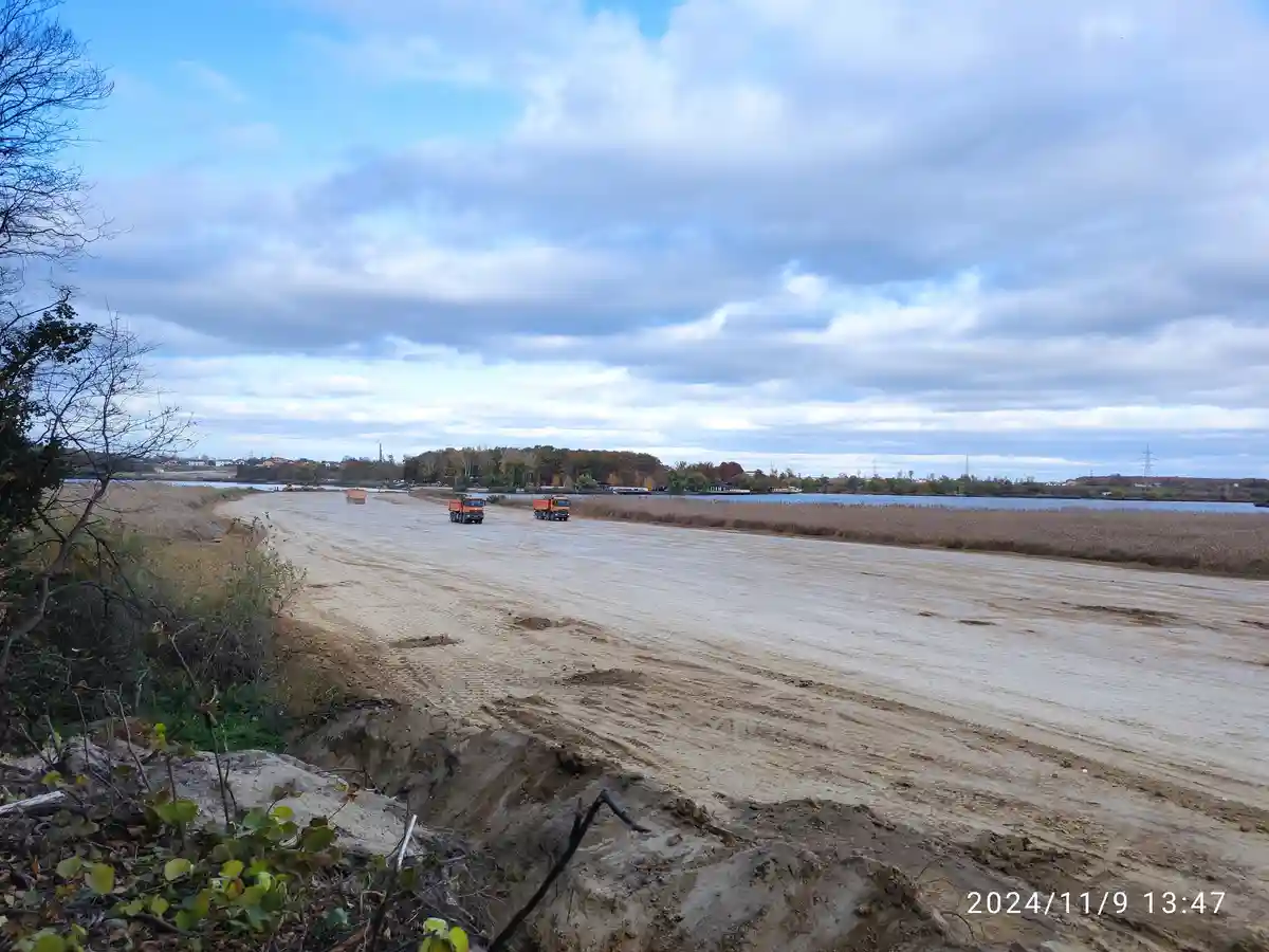 A wide, muddy construction road stretches beside a body of water under a cloudy sky, with three orange dump trucks driving…
