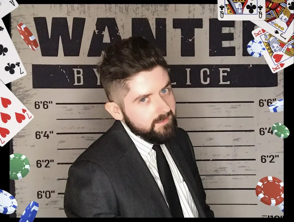 A man with a beard and blue eyes, wearing a suit and tie, poses in front of a 'WANTED BY POLICE' mugshot-style backdrop…