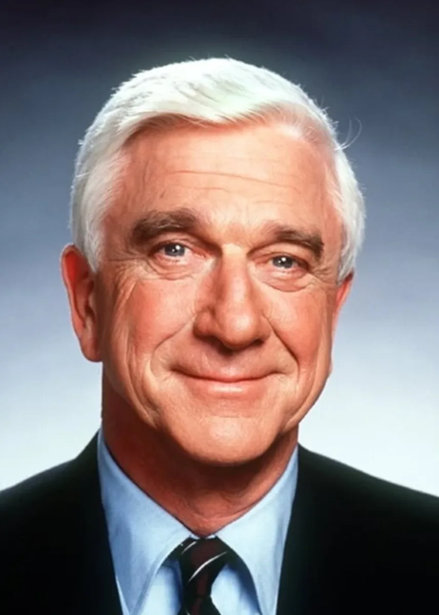 a professional headshot of an older white man with white hair, wearing a dark suit, light blue shirt, and dark tie, smiling…