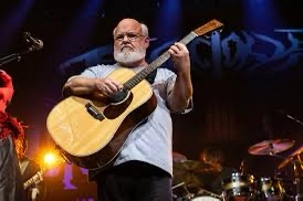 A man with a white beard and glasses plays an acoustic guitar on stage during a concert. Behind him, a band setup with drums…