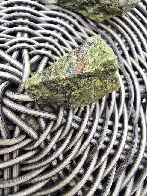A close-up photo shows a greenish, rough-textured rock resting on a woven metal or plastic basket with interlocking circular…