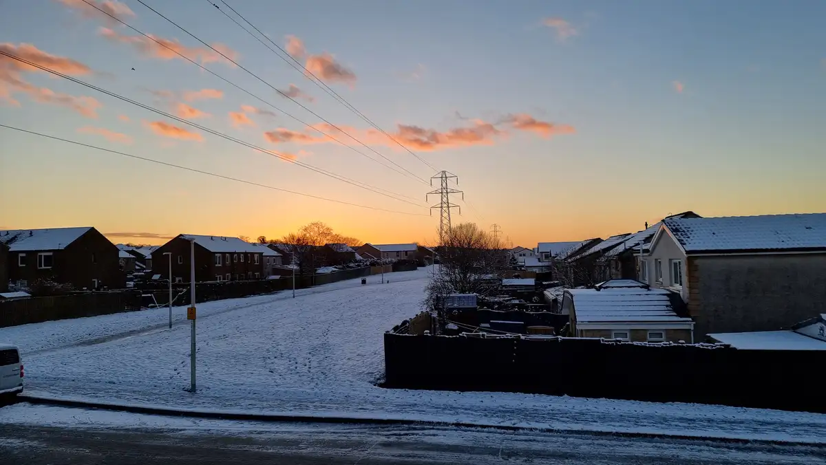 A snowy residential street at sunset, with houses covered in snow, power lines stretching across the sky, and a warm orange…