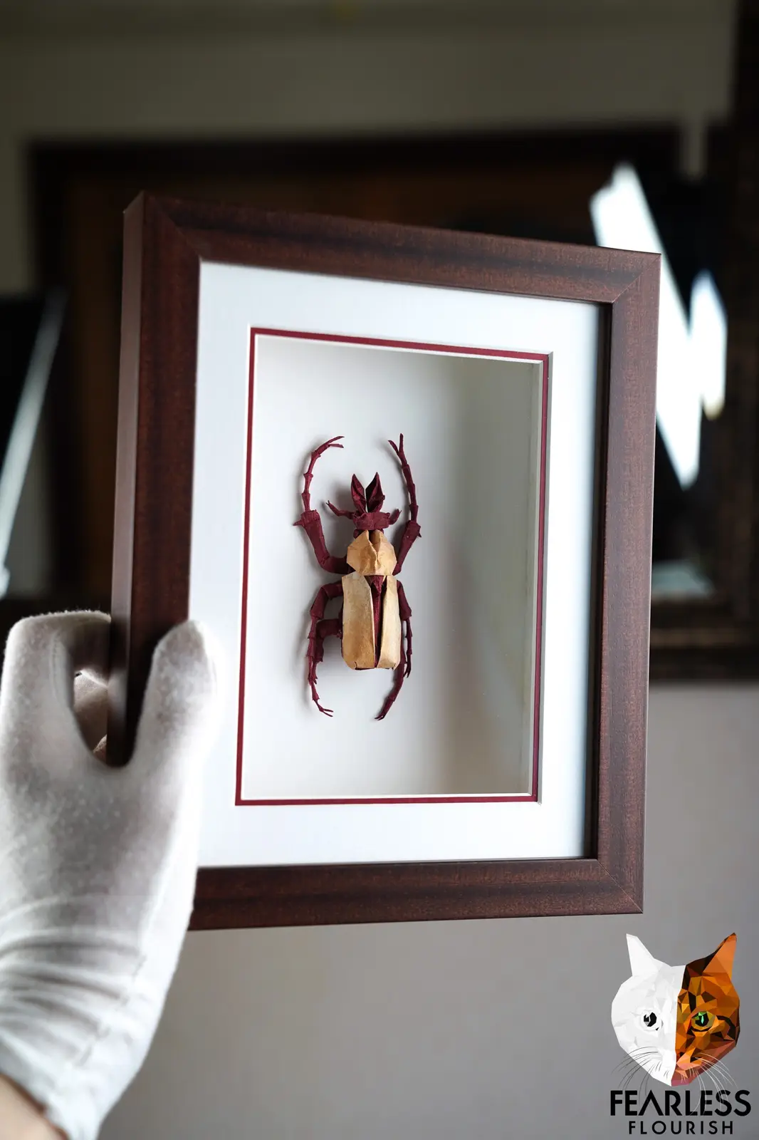 A hand in a white glove holds a dark wooden picture frame displaying a meticulously crafted origami beetle made of red and…
