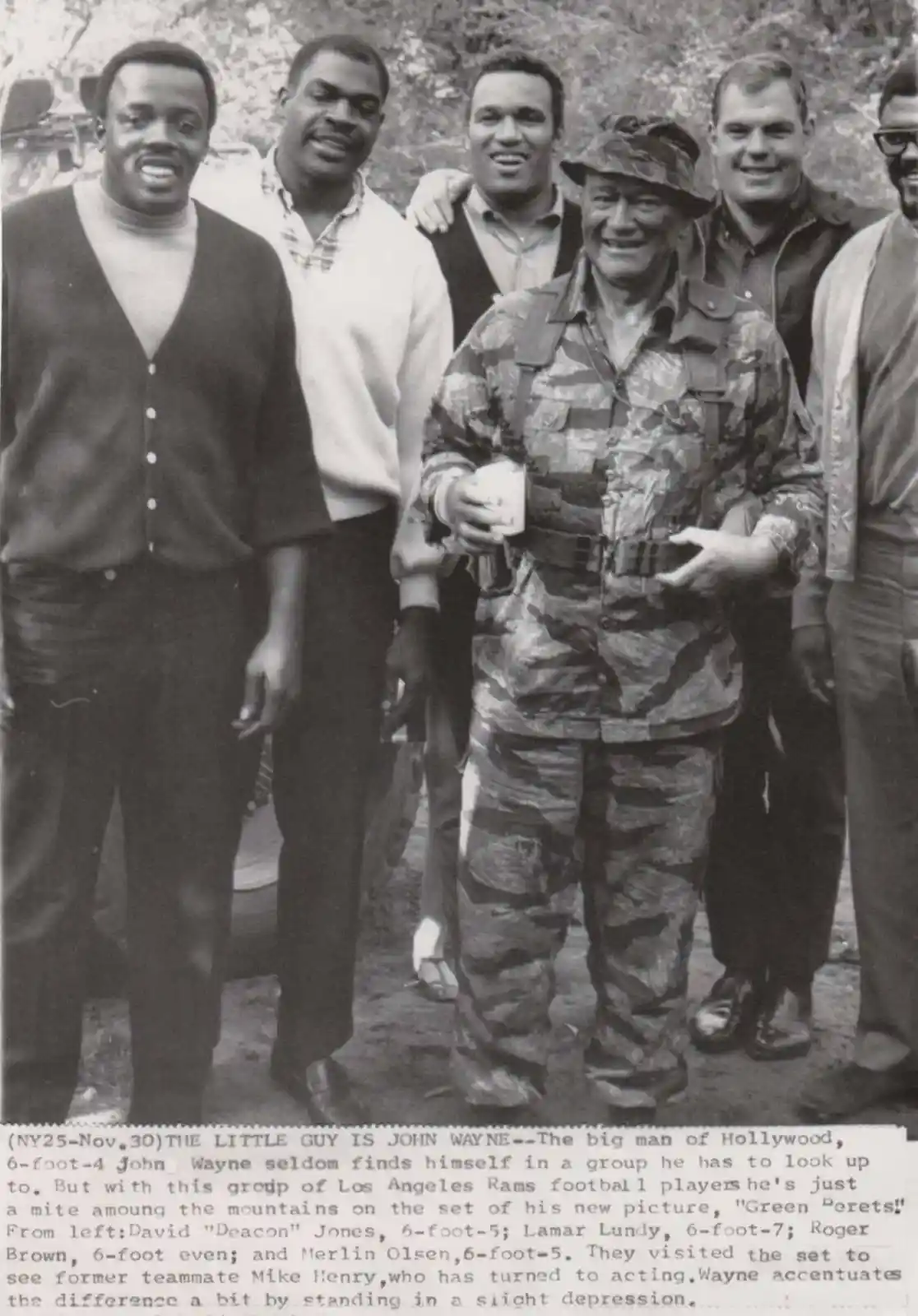 A black-and-white photo shows actor John Wayne, dressed in camouflage military gear and a hat, standing noticeably shorter…