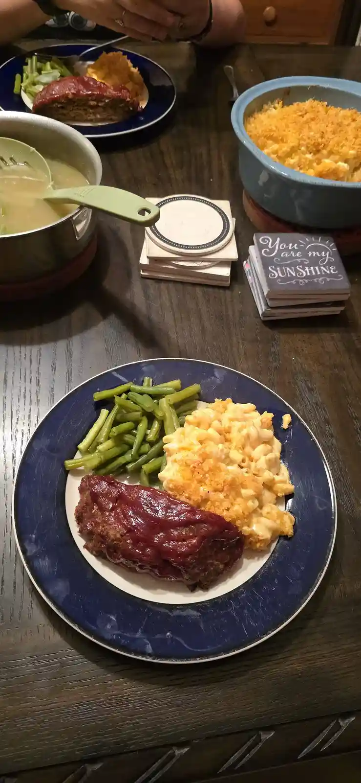 A close-up photo of a home-cooked meal on a dark wooden table, featuring a blue plate with meatloaf, macaroni and cheese…