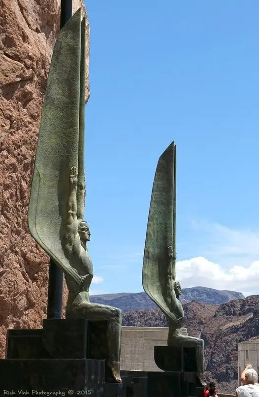 Two large bronze statues of seated figures with massive, wing-like blades held upright against a clear blue sky and rocky…