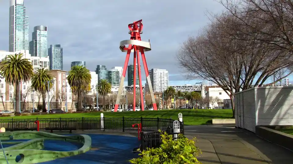 A daytime photo of a city park with a large red and silver abstract sculpture as the central focus, surrounded by green…