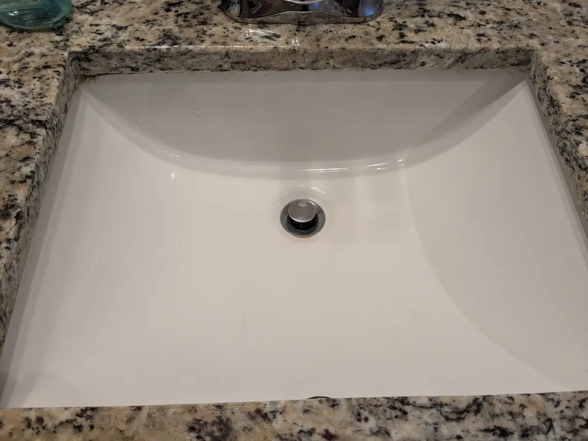 A top-down photo of a clean, white bathroom sink set into a speckled granite countertop, with a chrome faucet visible at the…