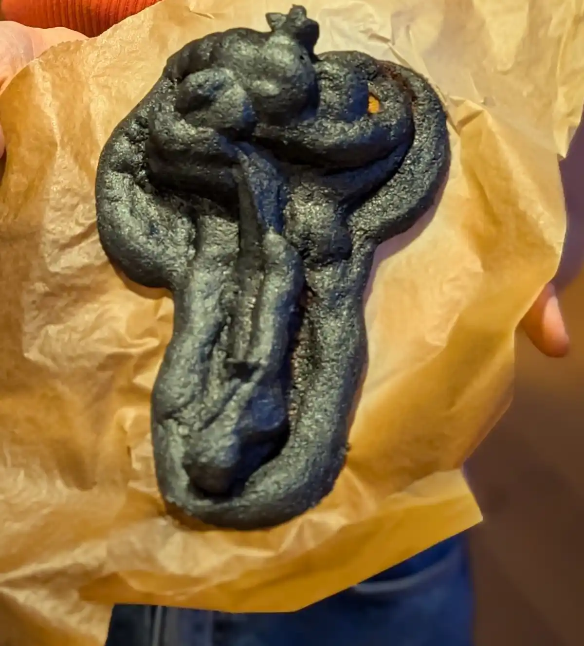 A close-up photo shows a dark, charred, and oddly shaped baked good held in someone's hands over brown paper. The item has a…