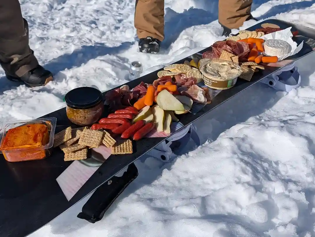 a snowboard is laid flat in deep snow and transformed into a charcuterie board, laden with meats, cheeses, crackers…