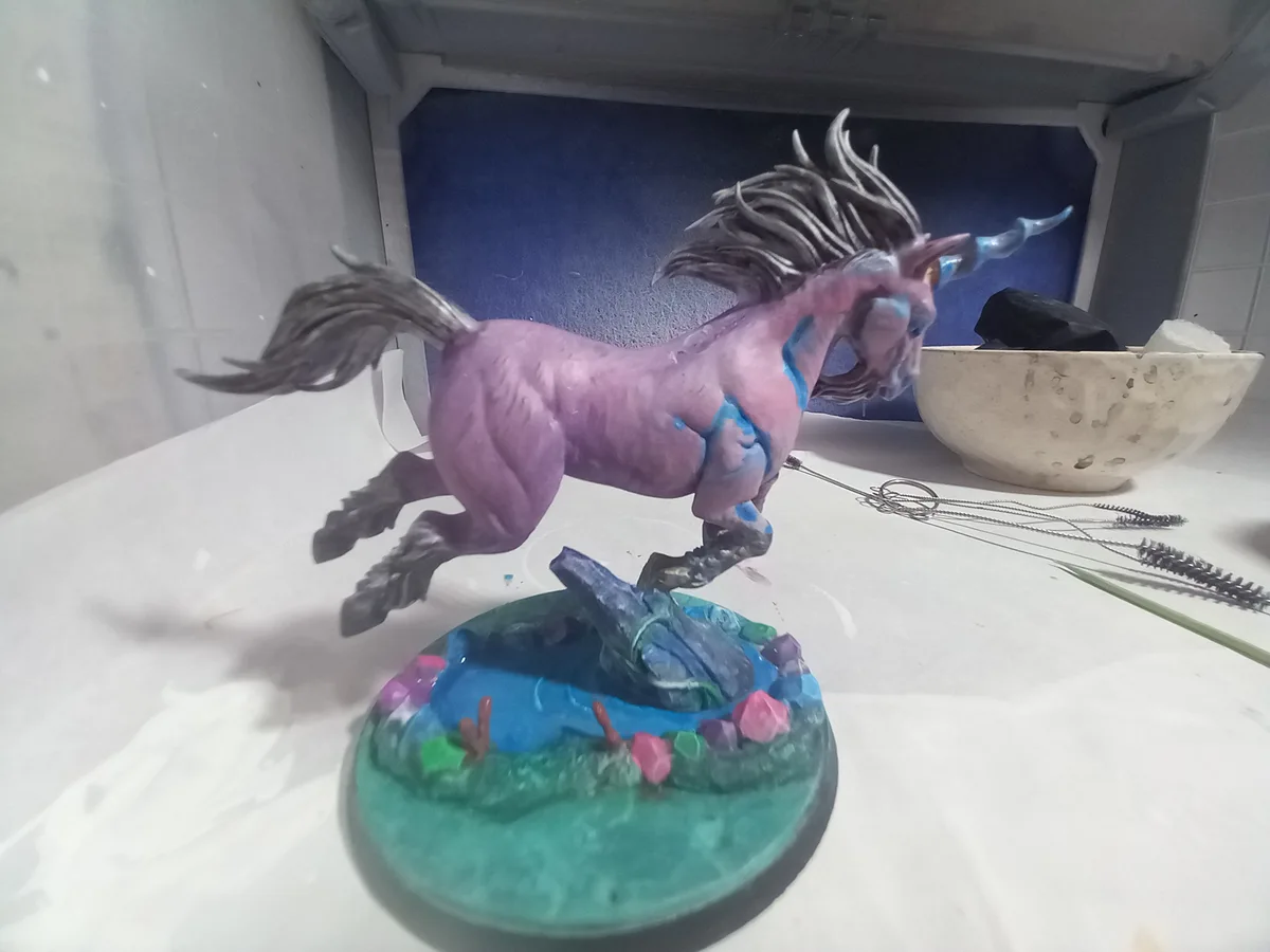 A partially painted miniature unicorn figurine, mostly purple with blue paint streaks, stands on a painted base resembling a…
