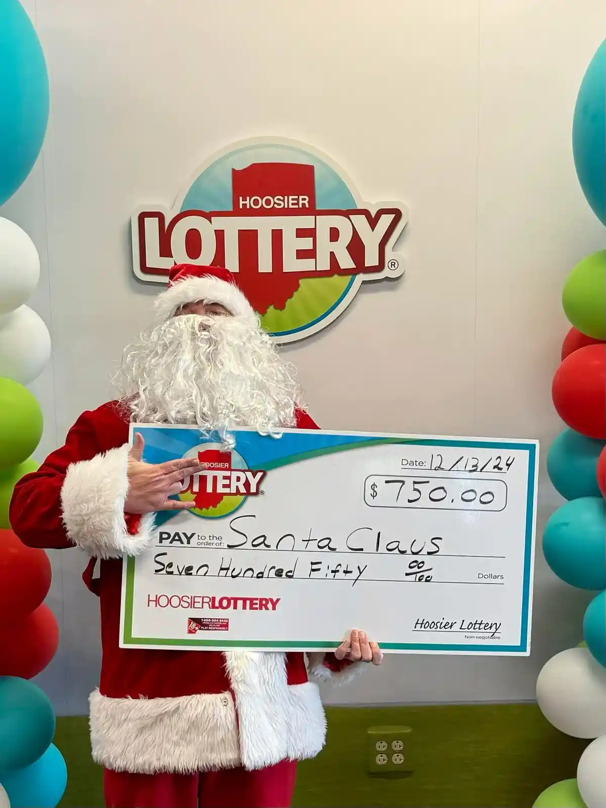 A person dressed as Santa Claus holds a large ceremonial check from the Hoosier Lottery for $750.00, payable to 'Santa…