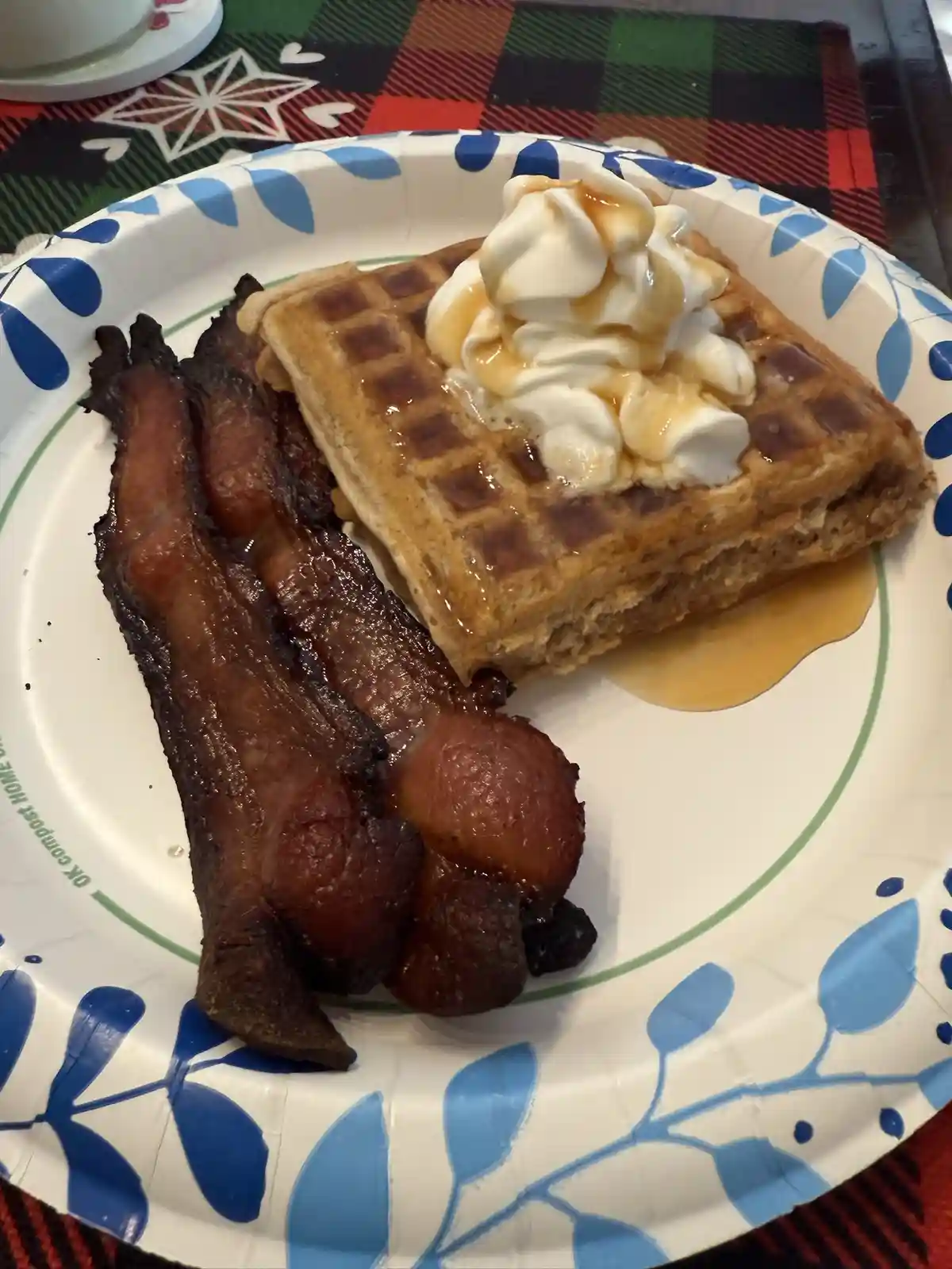 A close-up photo shows a plate of breakfast food: a square waffle topped with whipped cream and syrup, next to three strips…