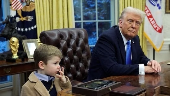 A young boy sits at a large wooden desk in an office resembling the Oval Office, looking thoughtful with his finger to his…