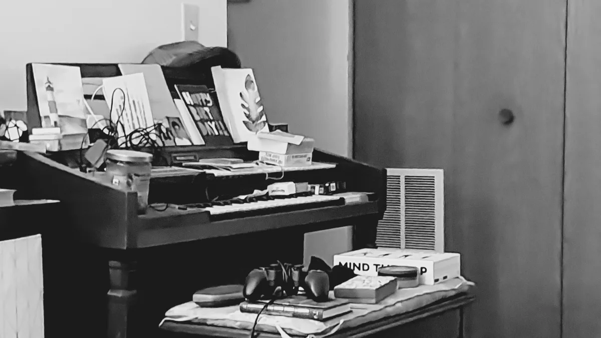 A black-and-white photo shows a cluttered piano with various items like greeting cards, a jar, and tangled wires on top…