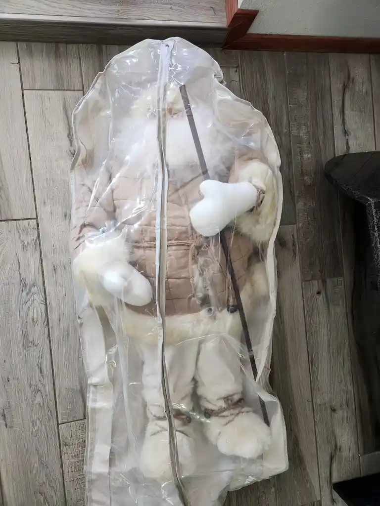 A large, plush Santa Claus figure is stored upright inside a clear plastic garment bag, standing on a wood-laminate floor…