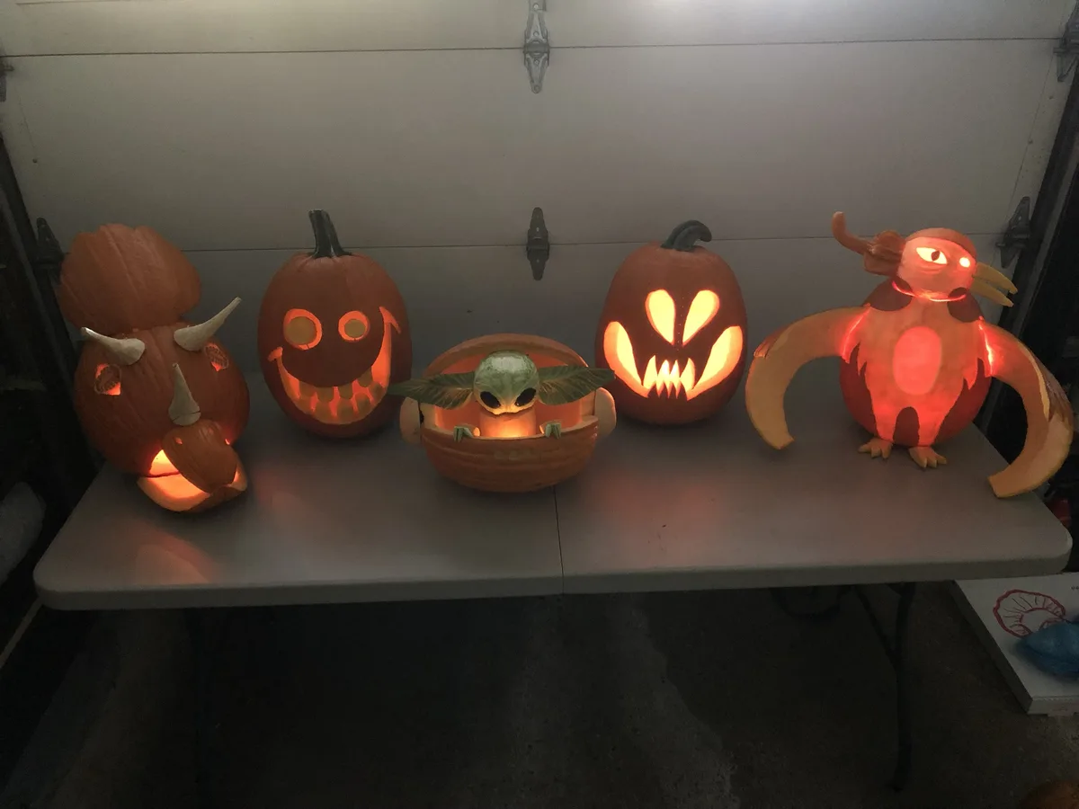 Five intricately carved, glowing pumpkins sit on a table, each shaped like a different character: a triceratops, a smiling…
