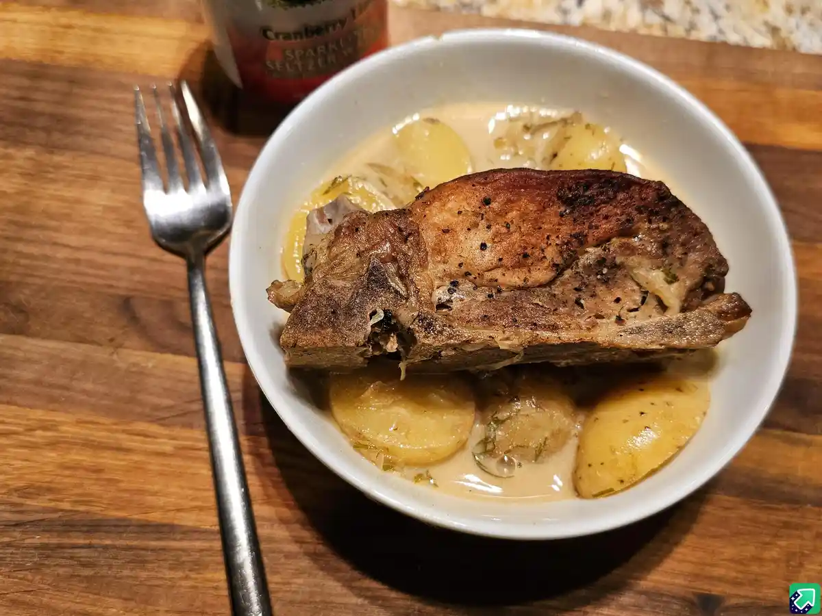 A close-up photo shows a white bowl on a wooden table containing a piece of seared meat with a bone, resting in a creamy…