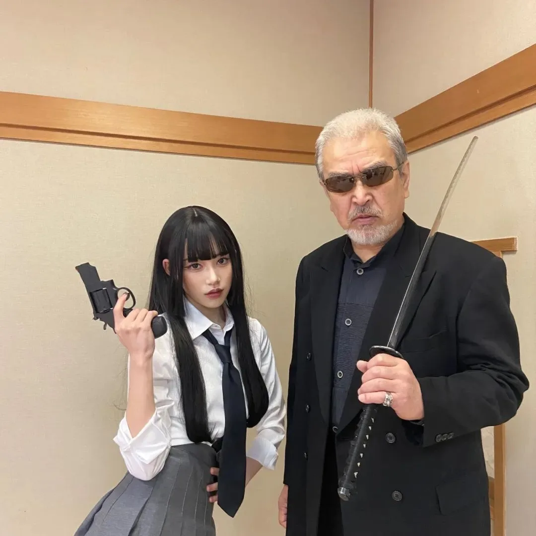 A young woman in a school uniform holds a revolver while standing beside an older man in a black suit holding a katana. Both…