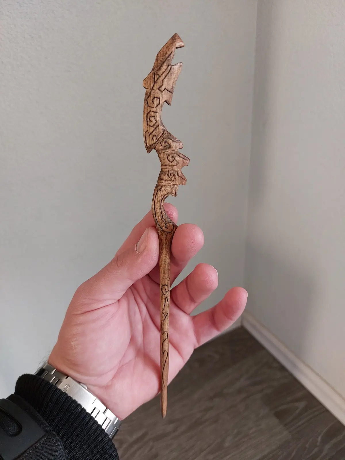 A hand holds a small, intricately carved wooden object resembling a wand with ornate patterns and a pointed tip, against a…