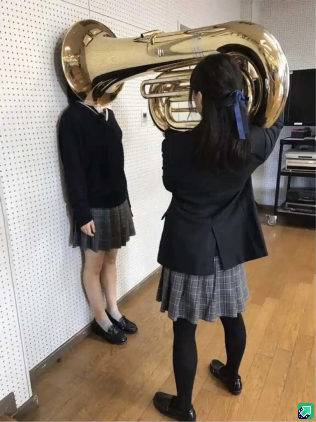 Two girls in school uniforms stand in a room; one leans against a wall with a large tuba placed over her head, while the…