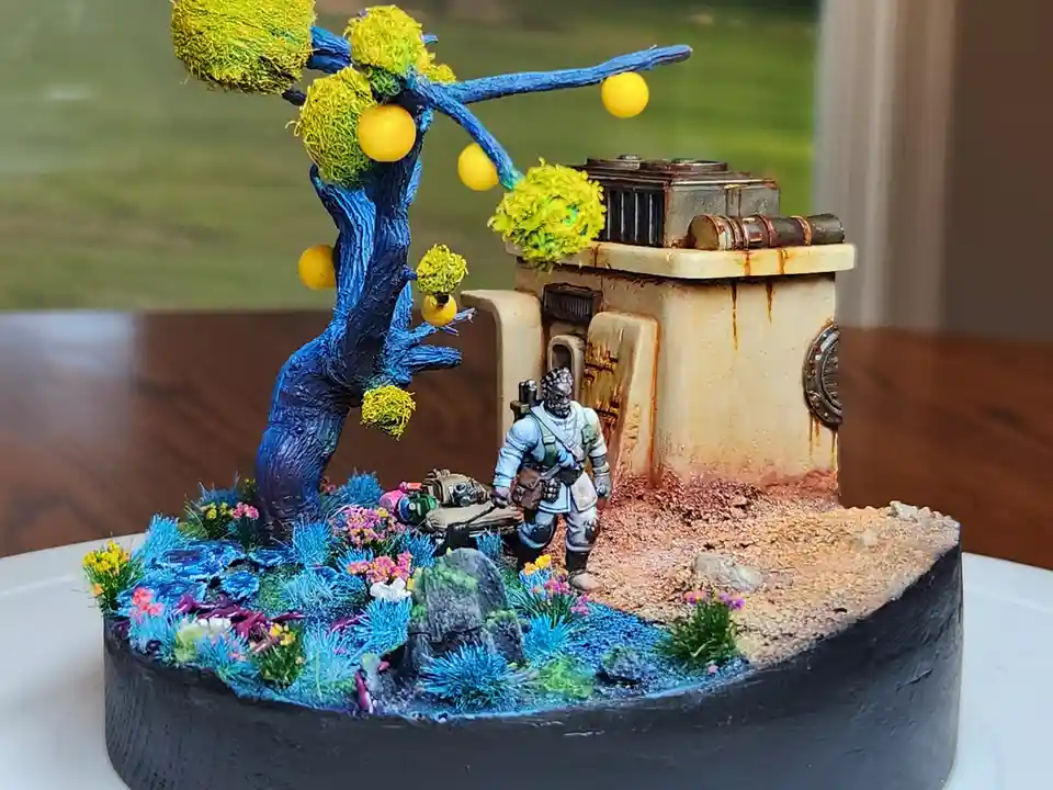 A miniature diorama features a painted blue tree with yellow spherical fruit and fuzzy green foliage, standing beside a…