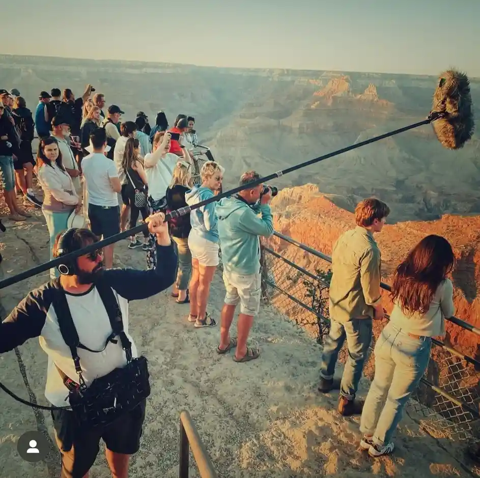 A man with headphones and a boom microphone extended over a crowd of tourists at the Grand Canyon, as if filming them. The…