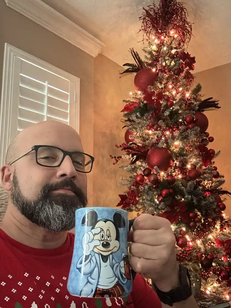 A man with glasses and a beard, wearing a red ugly christmas sweater, holds up a mug featuring a cartoon mickey mouse…
