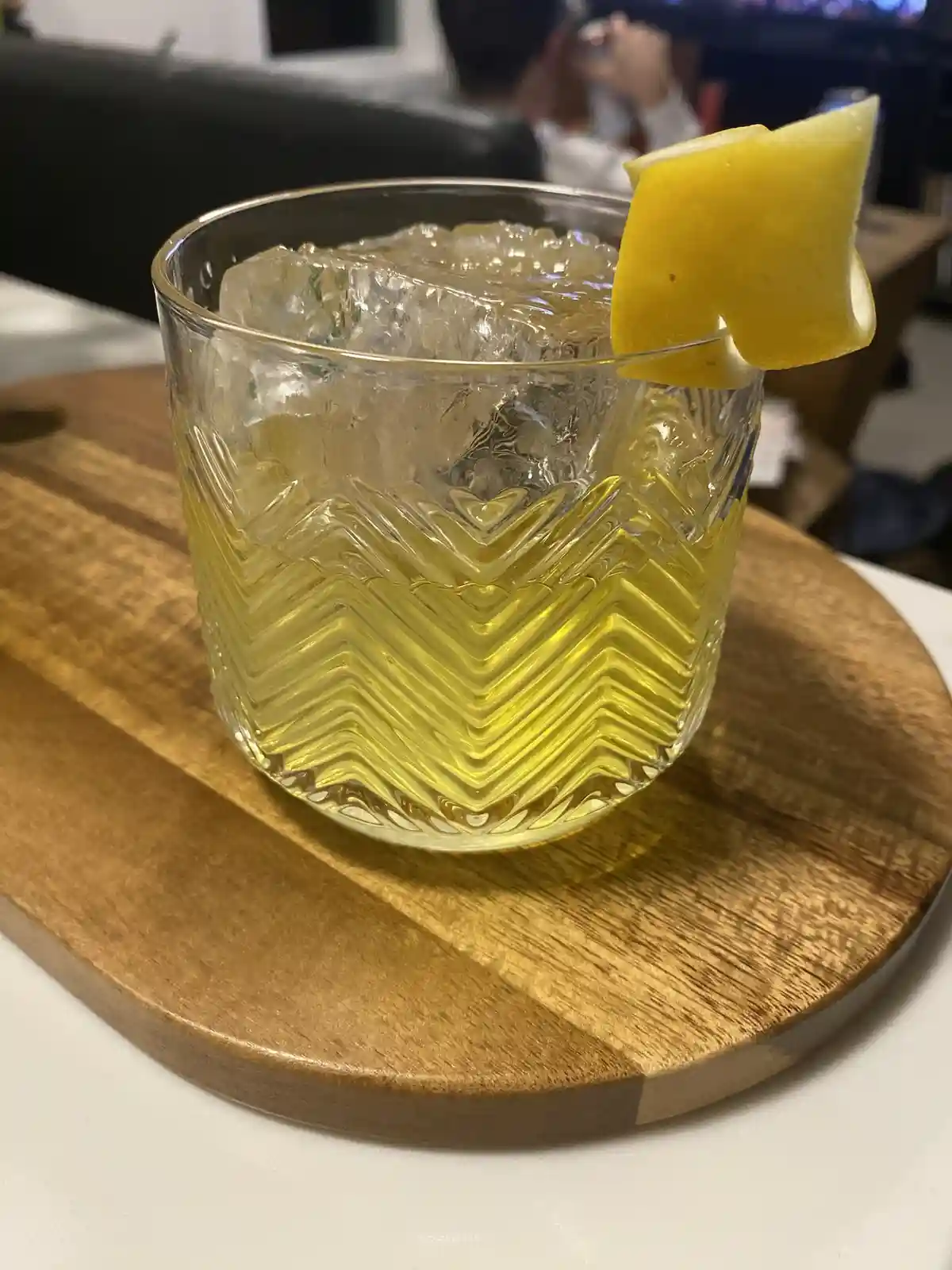 A close-up photo shows a cocktail in a textured glass with a zigzag pattern, filled with ice and a yellow liquid, garnished…