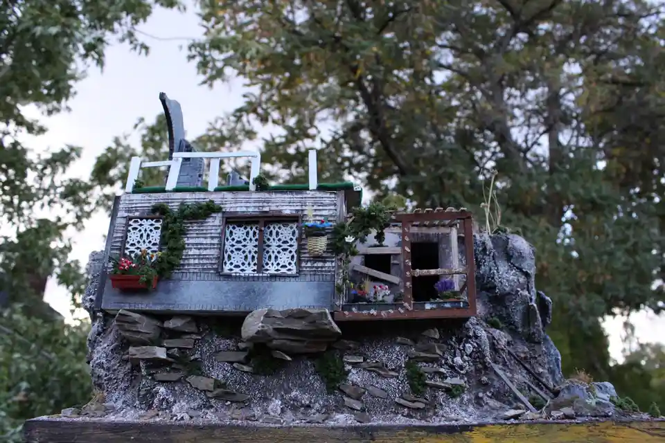 A miniature model of a rustic, weathered house perched precariously on a rocky cliffside, adorned with tiny flowers, ivy…