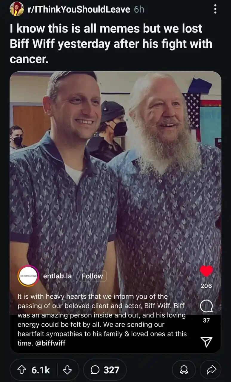 The image shows two men smiling for a photo; the man on the right has a long white beard and is wearing a patterned shirt…