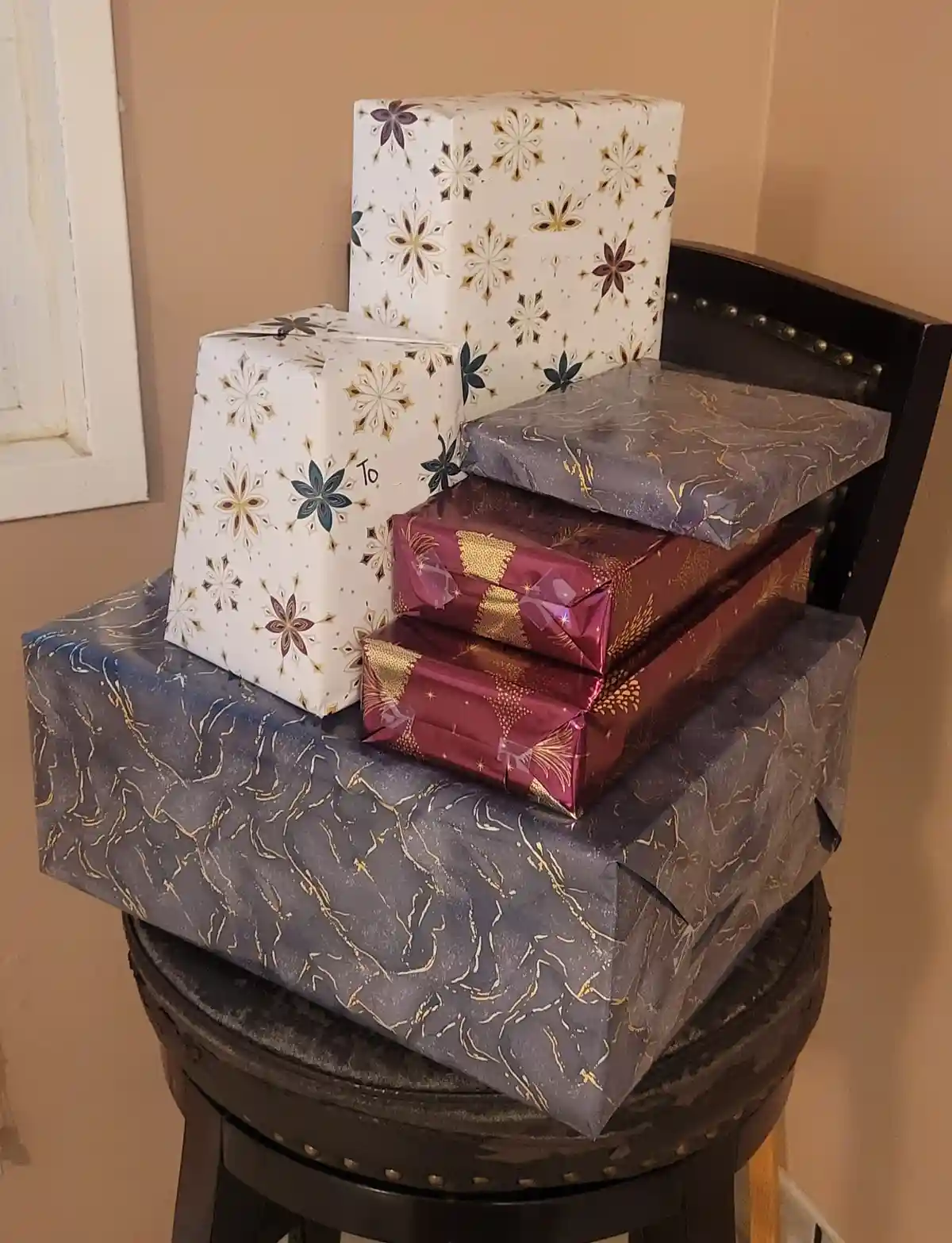 A stack of several Christmas presents wrapped in festive paper sits precariously on a black bar stool. One white gift box…