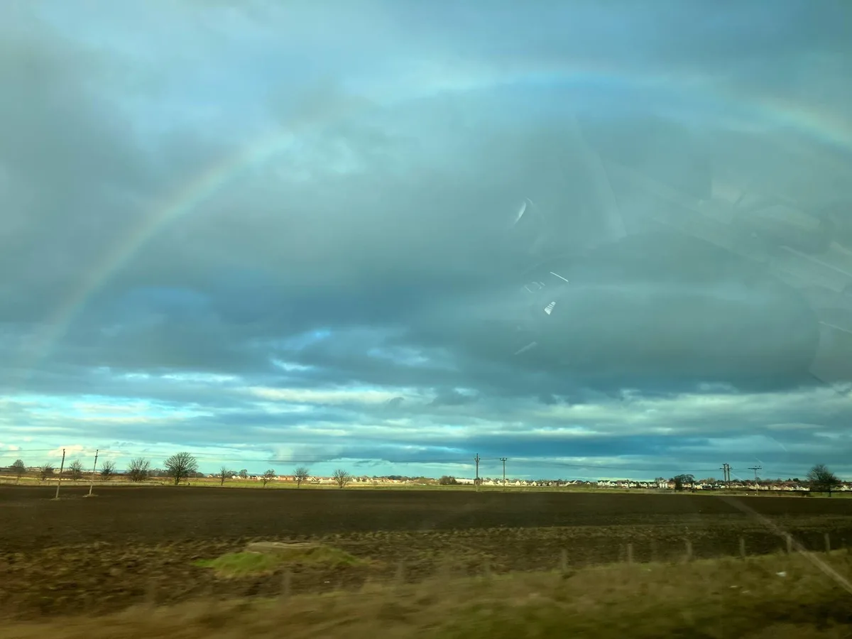 A photograph taken from inside a moving vehicle shows a faint rainbow arching across a cloudy sky over a rural landscape…