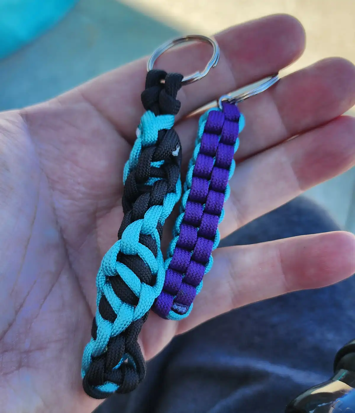 A hand holds two handmade paracord keychains, one braided with black and teal cords, the other with purple and teal cords…
