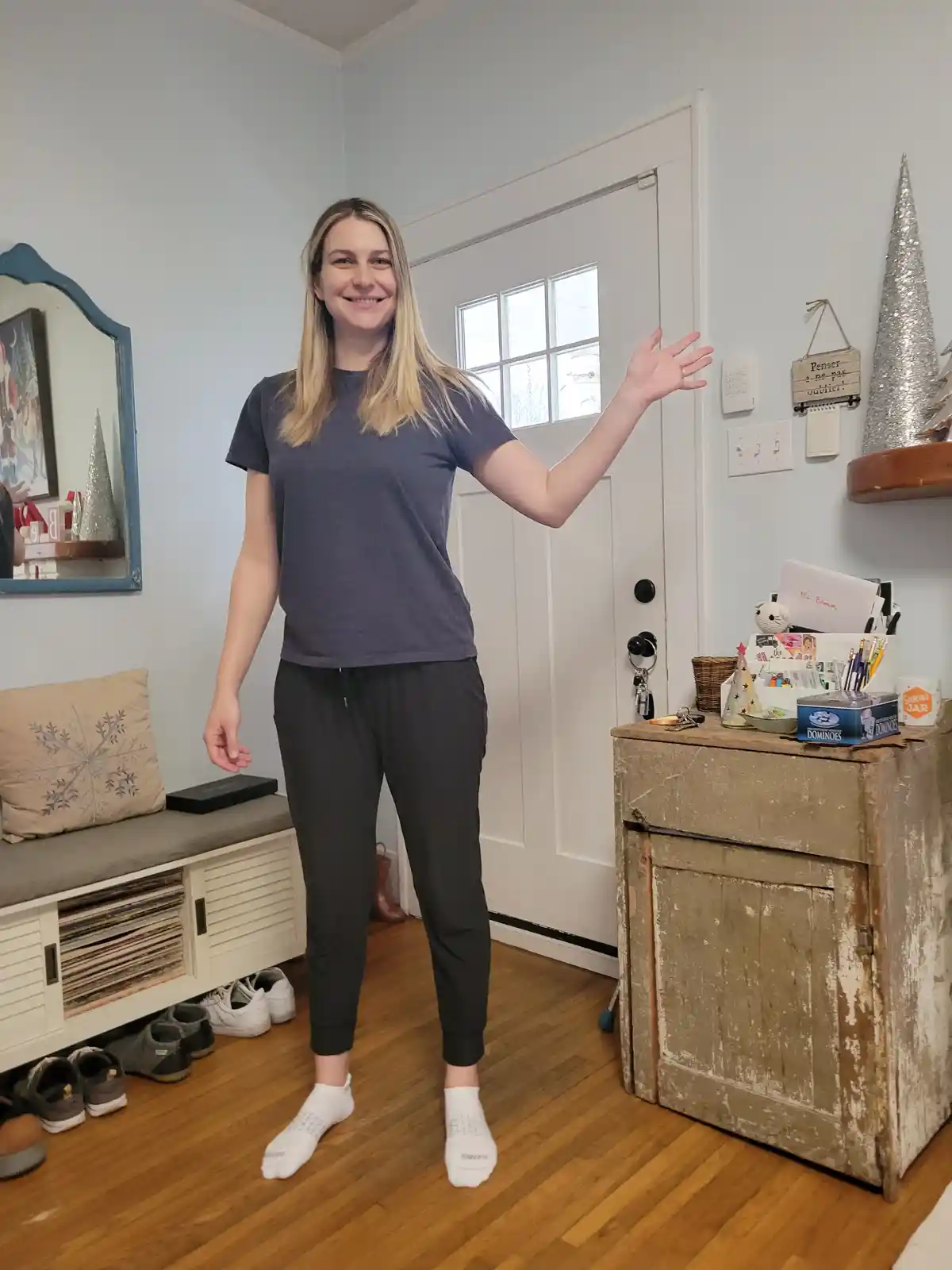 A woman in a grey t-shirt and black joggers stands in a home entryway, smiling and gesturing toward a wall with a thermostat…