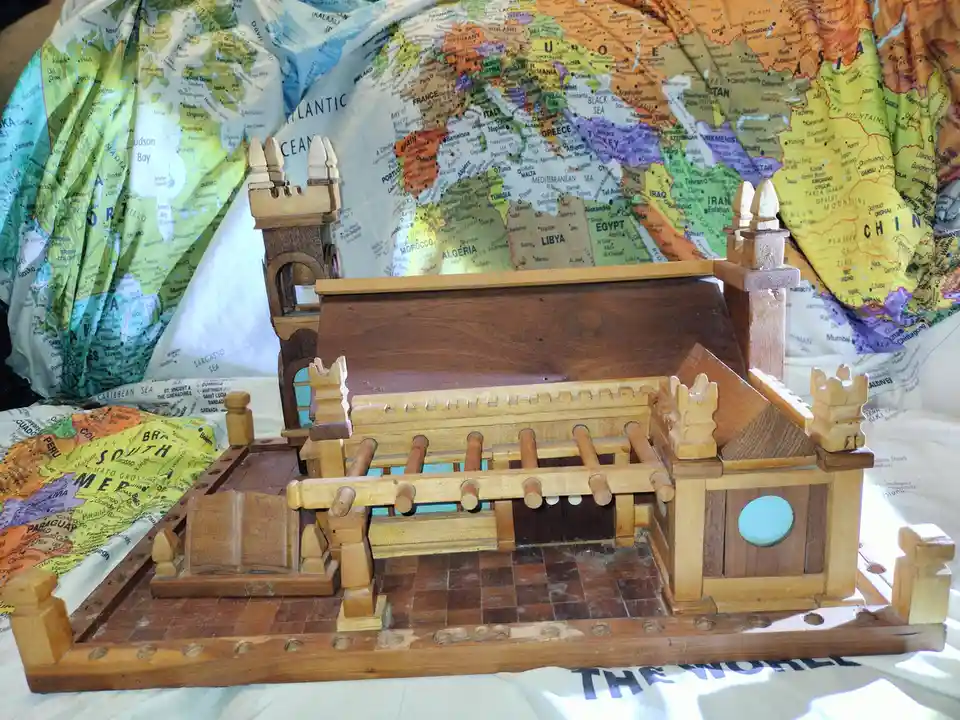 A wooden model of a church or cathedral with a checkered floor, open book, and crenellated towers sits atop a world map. The…
