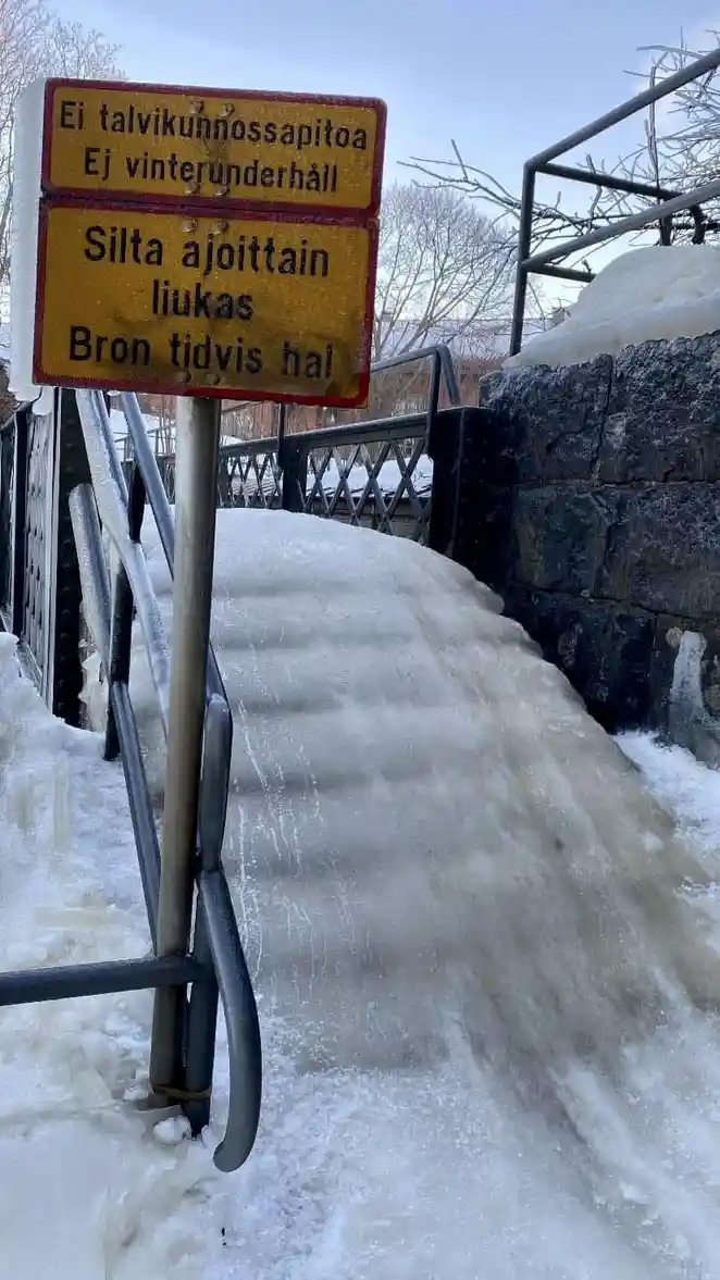 A yellow warning sign with red borders stands beside a snow-covered bridge ramp, where ice has formed a thick, slippery…