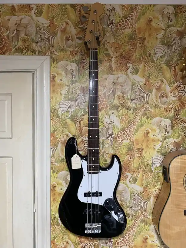 A black Fender Jazz Bass guitar hangs against a wall covered in dense, colorful wallpaper featuring numerous African animals…