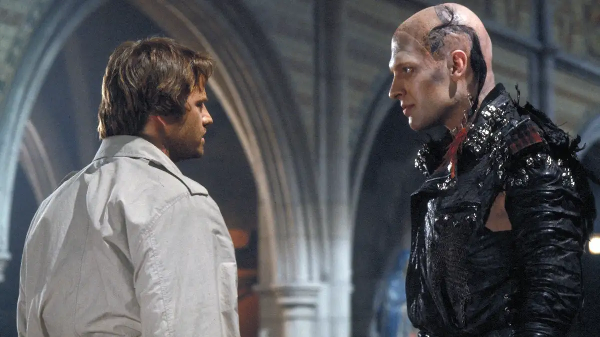 two men face each other in a gothic-style setting; one wears a plain light-colored shirt, the other has a shaved head with a…