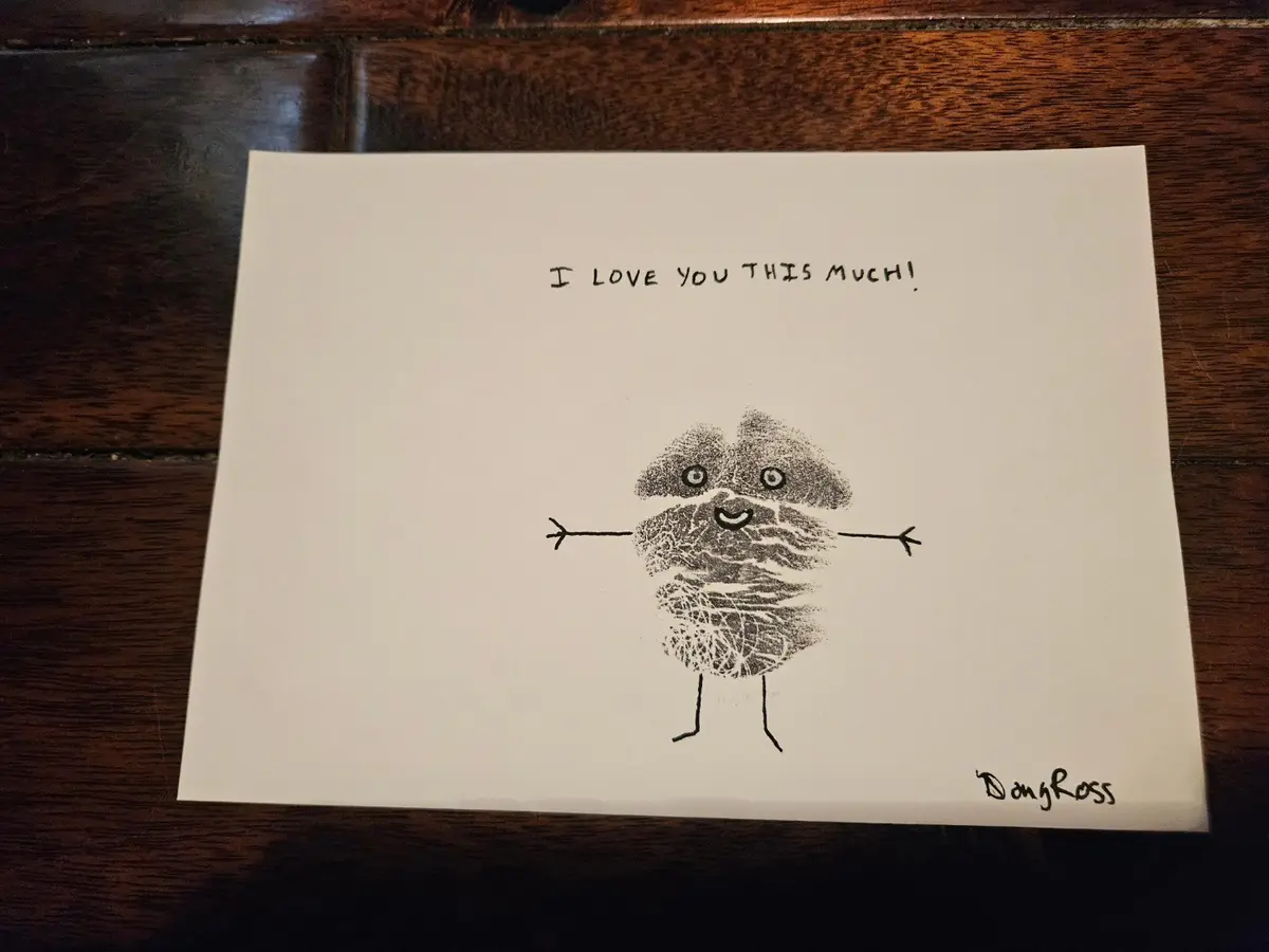 A handmade card on white paper shows a childlike drawing of a smiling, stick-figure-like creature made from a smudged…