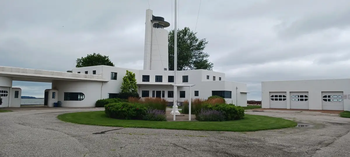 A wide shot of a white, modernist-style building with a prominent tower and flagpole, surrounded by gravel, grass, and…